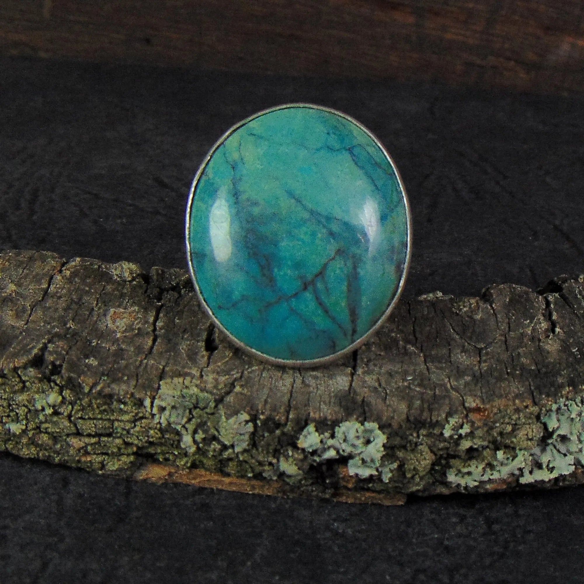 Pierścionek z błękitnym turkusem od EPIR Art Jewellery&Gemstone to polerowany turkusowy kamień oprawiony w elegancką srebrną obrączkę, efektownie prezentującą się na teksturowanym, pokrytym porostami kawałku drewna na rustykalnym, ciemnym tle.